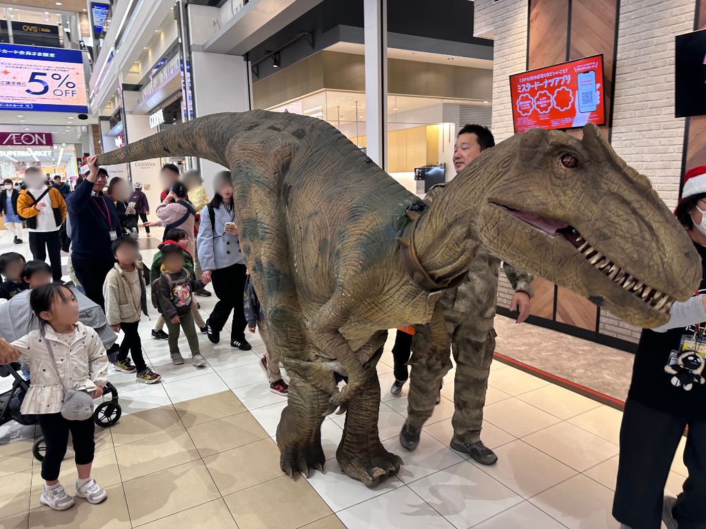 商業施設内を練り歩くリアルな恐竜のクローズアップ。子どもたちが見上げる迫力のある姿